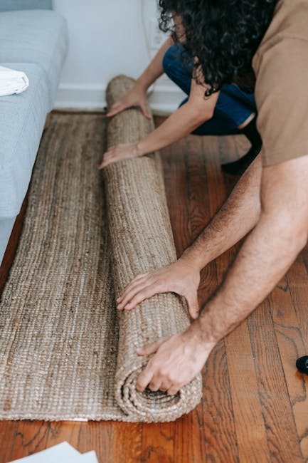 A person is carefully unrolling a beige and brown patterned carpet onto a polished wooden floor inside a residential living room. The carpet appears clean and well-maintained, with a soft texture visi