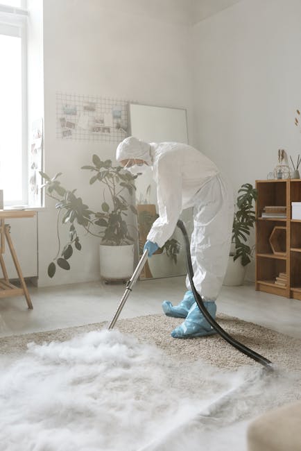 A professional cleaner dressed in a white protective suit, gloves, and a face mask is performing surface cleaning on a plush beige carpet in a well-lit, modern living room. The cleaner is using a vacu