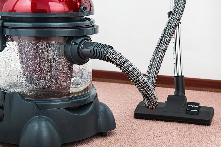 A person dressed in full protective cleaning gear, including a white suit, mask, gloves, and shoe covers, performs carpet cleaning in a well-lit, modern living room. They are using a steam cleaning ma
