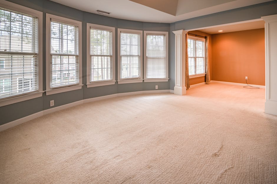 A spacious residential living room with a large bay window featuring multiple white-framed, double-glazed panes and white wooden blinds, allowing natural light to fill the area. The room has light gre