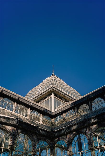 Exterior view of a historic glass-domed conservatory with ornate iron framework and large arched windows, featuring intricate decorative detailing on the metalwork. The building appears to be made of 
